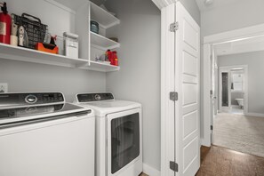 Une buanderie équipée de deux machines à laver et de deux sèche-linge, d’étagères contenant des produits de nettoyage et d’une porte donnant accès à une salle de bain.