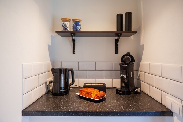 Una cocina con encimera negra, baldosas blancas, un tostador, una cafetera y un plato con comida.