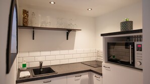A modern kitchen with white cabinets, a black countertop, a built-in microwave, and a shelf with glassware and a plant. - Garden View (Double or Twin) (Overschild)