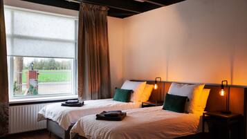A hotel room with two beds, a large window with curtains, a radiator, and a nightstand with a lamp.