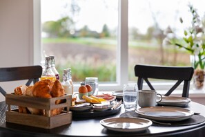 Una mesa de desayuno con croissants, huevos y botellas de jugo, frente a un paisaje verde.