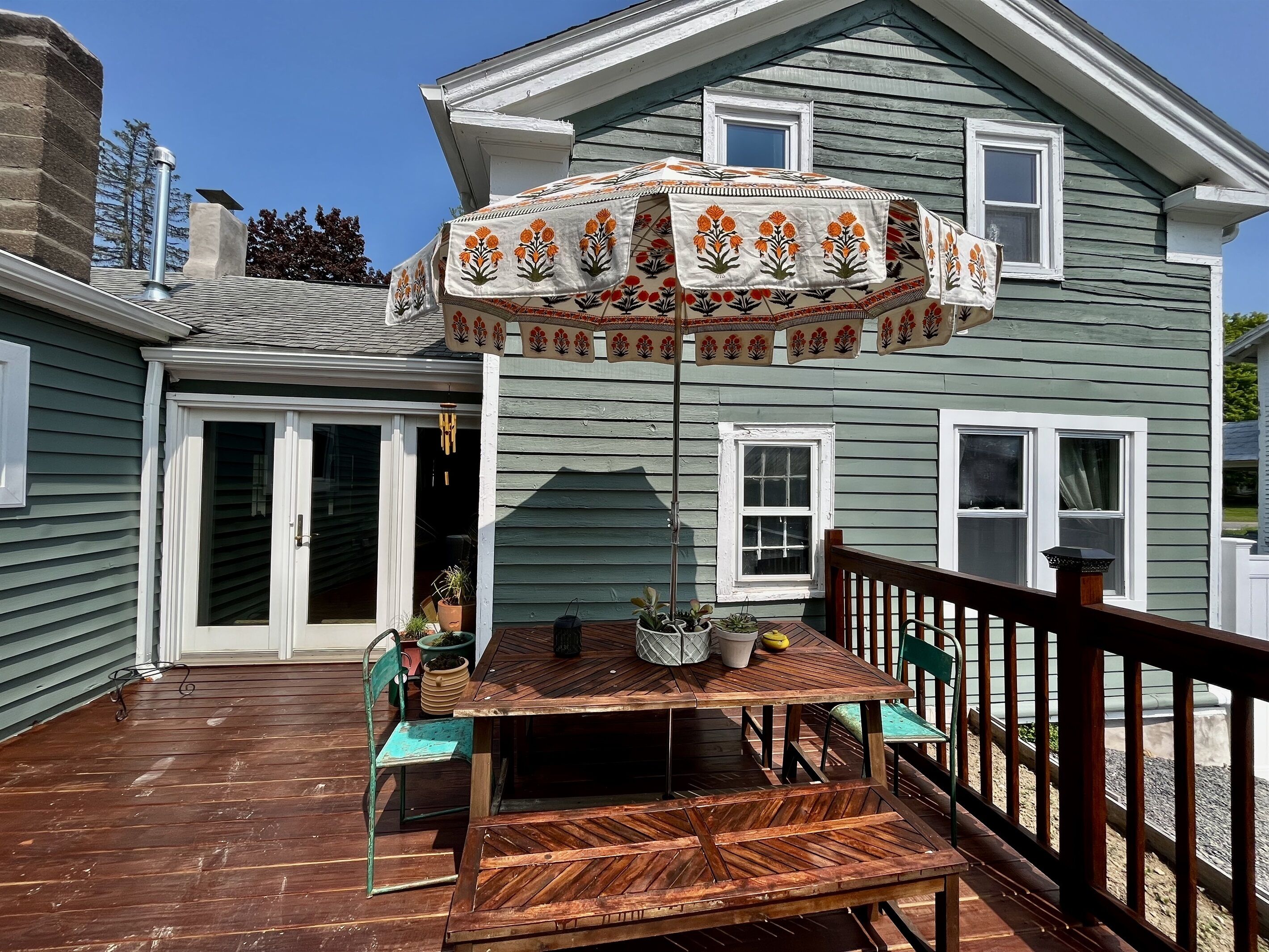 A wooden deck with a table and chairs, a covered patio area with a decorative umbrella, and a house with a green exterior and white trim.