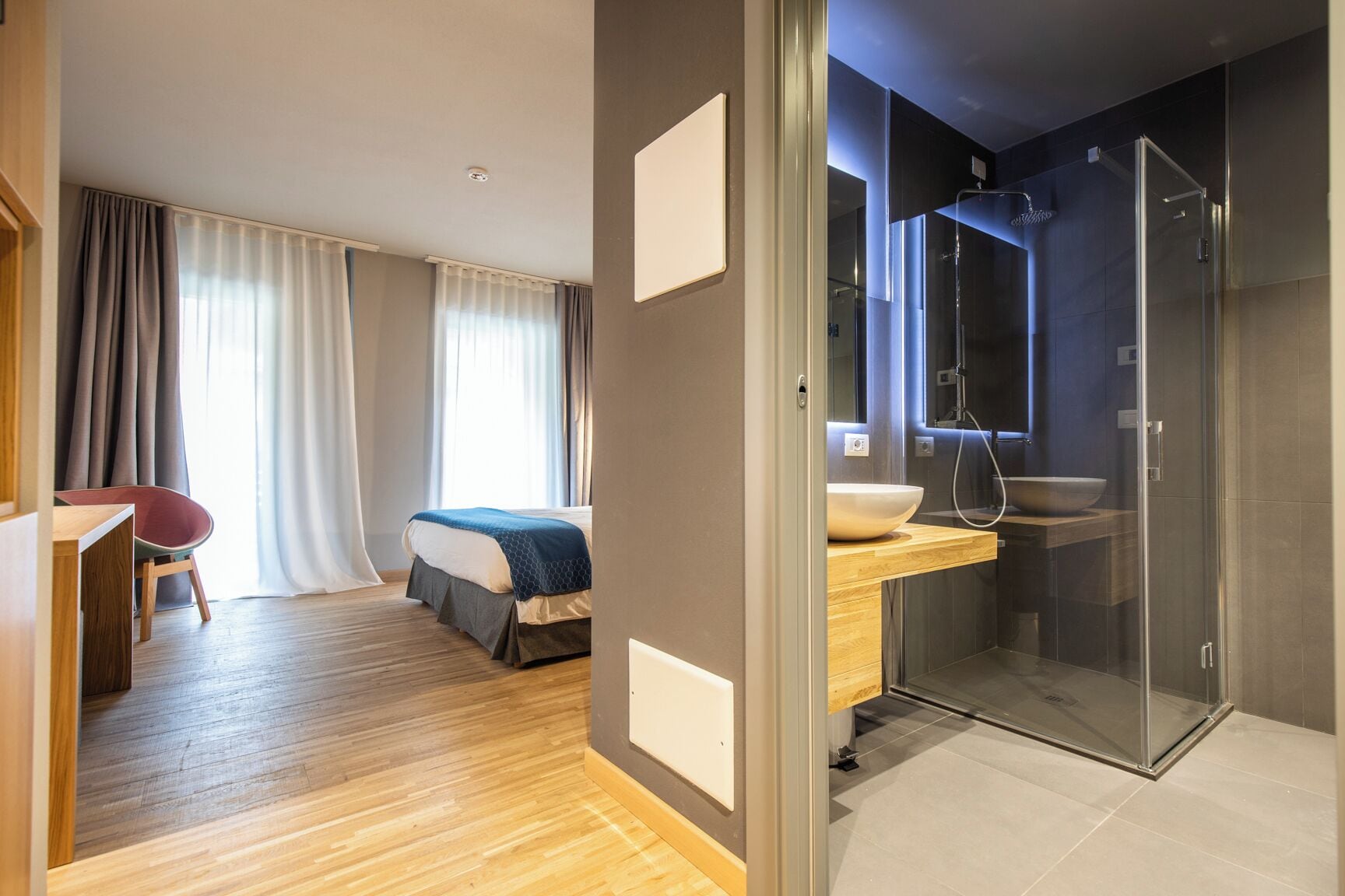 Ein modernes Hotelzimmer mit Holzboden, einem Bett mit blauer und weißer Bettdecke, einem Schreibtisch mit Stuhl sowie einem Badezimmer mit Duschkabine und großem Waschbecken.