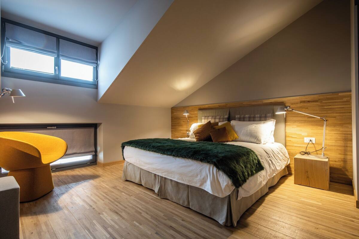 A modern bedroom with a large bed, a wooden headboard, a window with blinds, and a yellow chair.