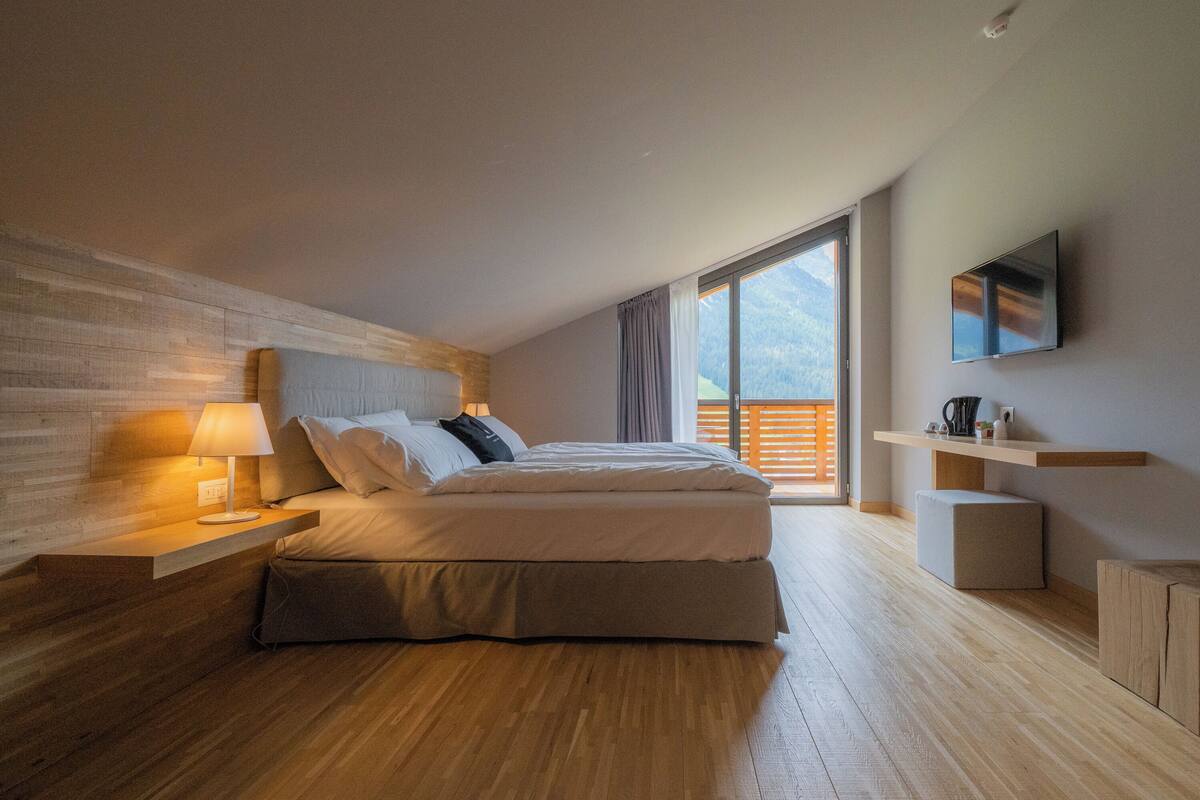 A modern bedroom with a wooden floor, a bed with white bedding, a nightstand with a lamp, a TV on the wall, and a balcony with a view of the mountains.