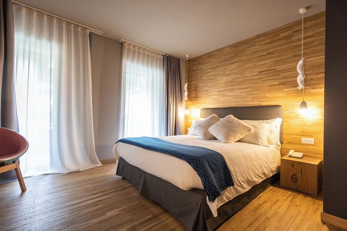 A modern hotel room with a wooden headboard, a large bed with white and dark blue bedding, a wooden nightstand with a phone, and a chair.