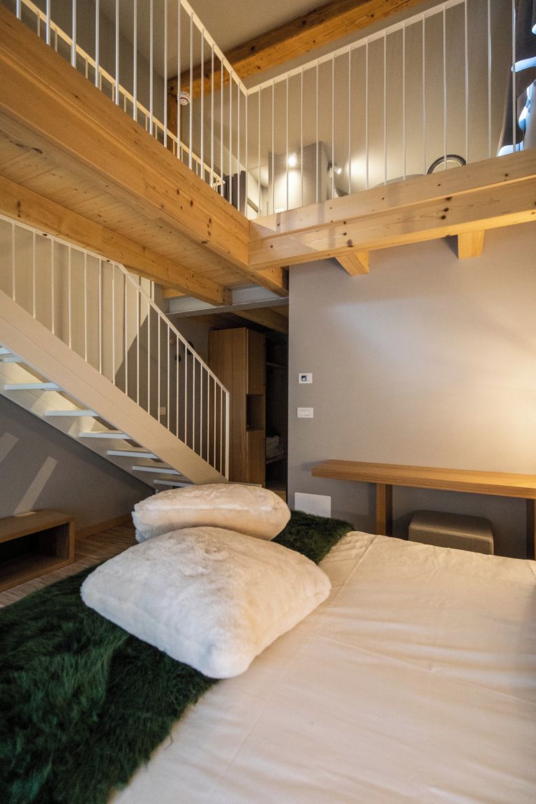 A bedroom with a bed, pillows, a green rug, a wooden bench, and a staircase with wooden railings.