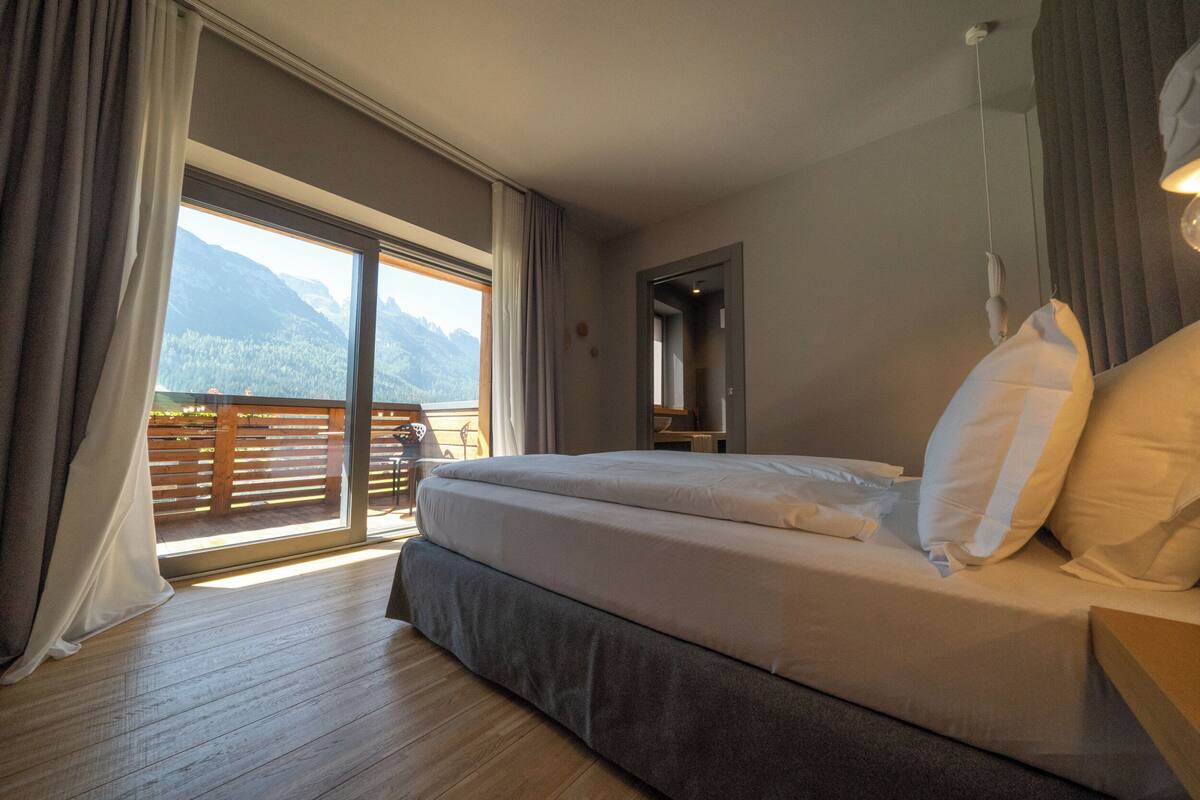A bedroom with a large bed, a balcony with a view of mountains, and a wooden floor.