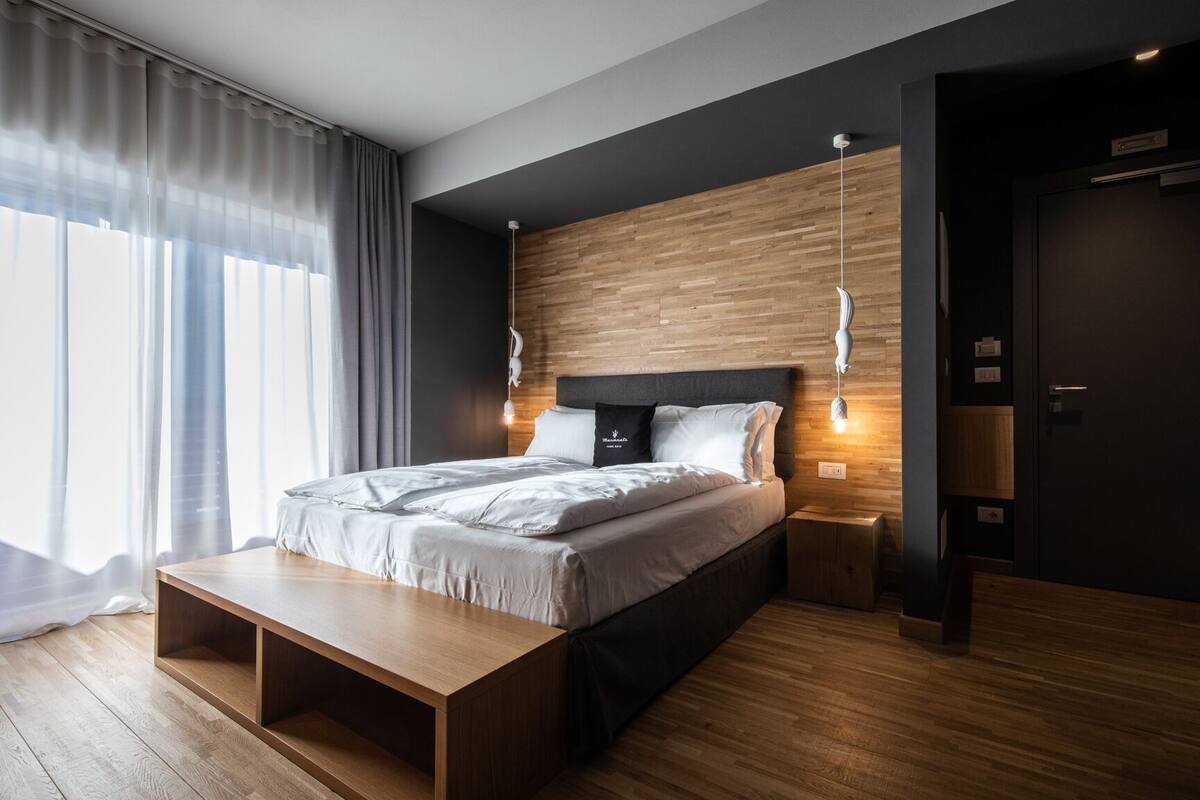A modern bedroom with a wooden headboard, a wooden bench at the foot of the bed, and a bedside table with a lamp.