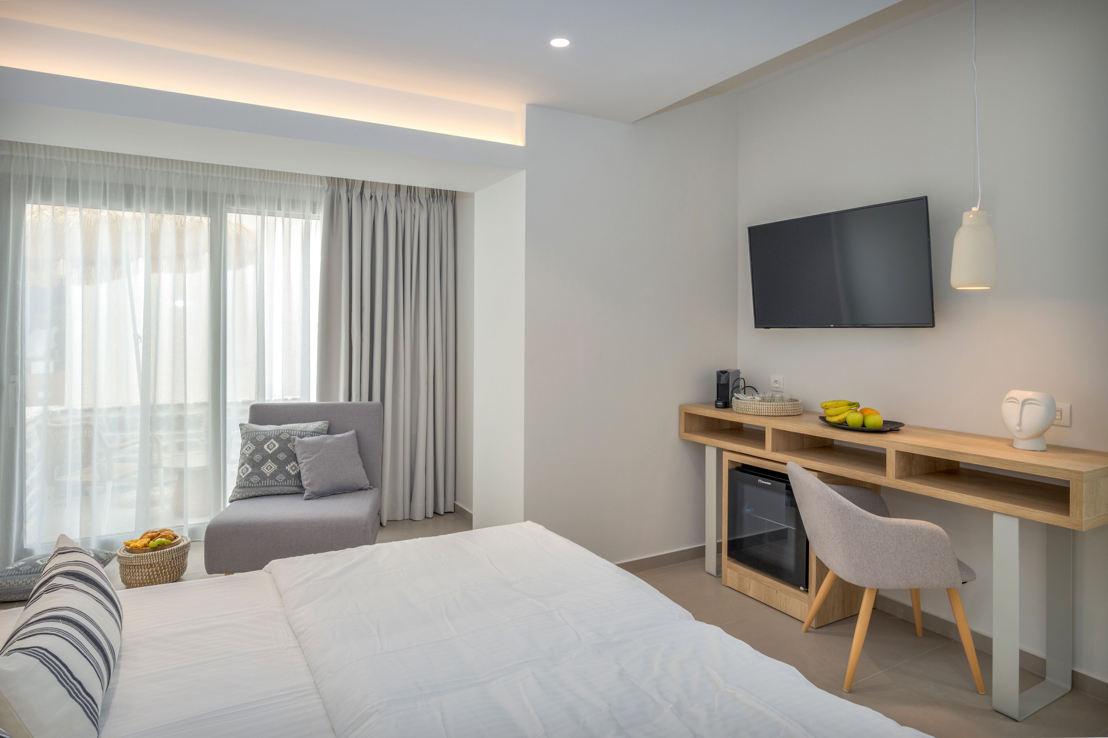 A modern hotel room with a large bed, a desk with a computer, a TV, and a small refrigerator.