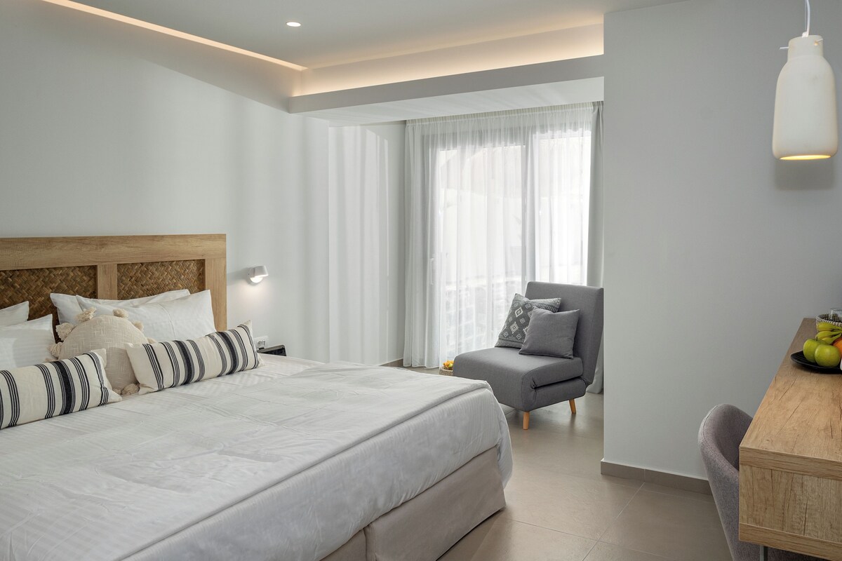 Ein modernes Hotelzimmer mit einem großen Bett, einem grauen Sessel, einem Holztisch und einem großen Fenster mit durchsichtigen Vorhängen.