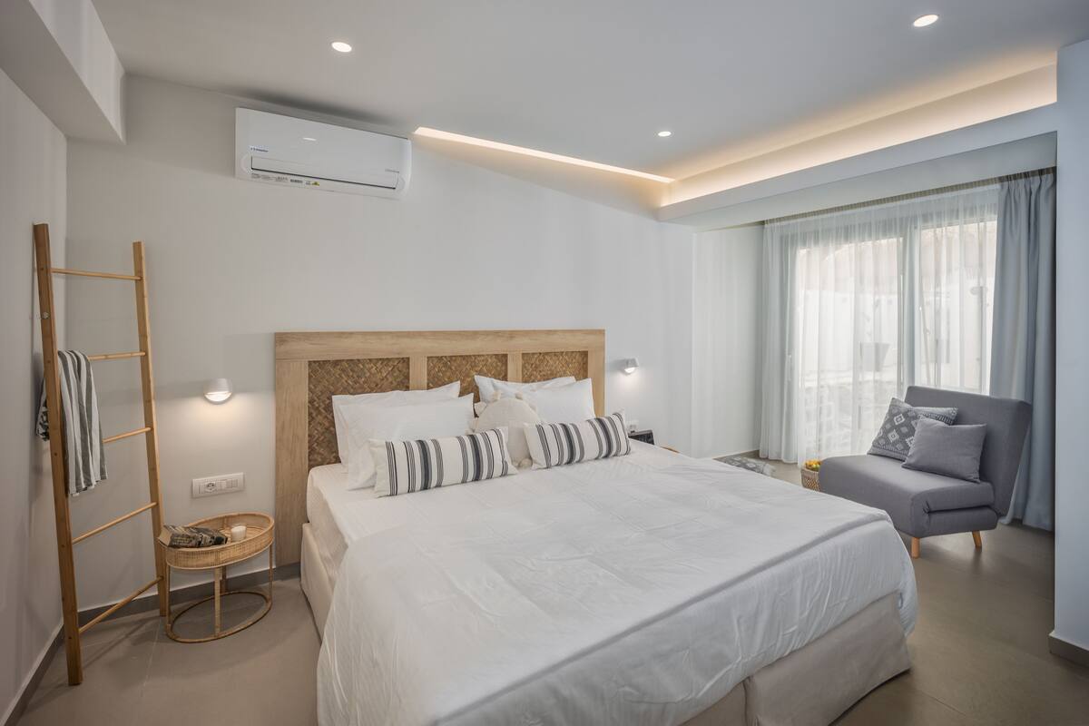 A modern hotel room with a large bed, a grey armchair, a small side table, and a window with sheer curtains.