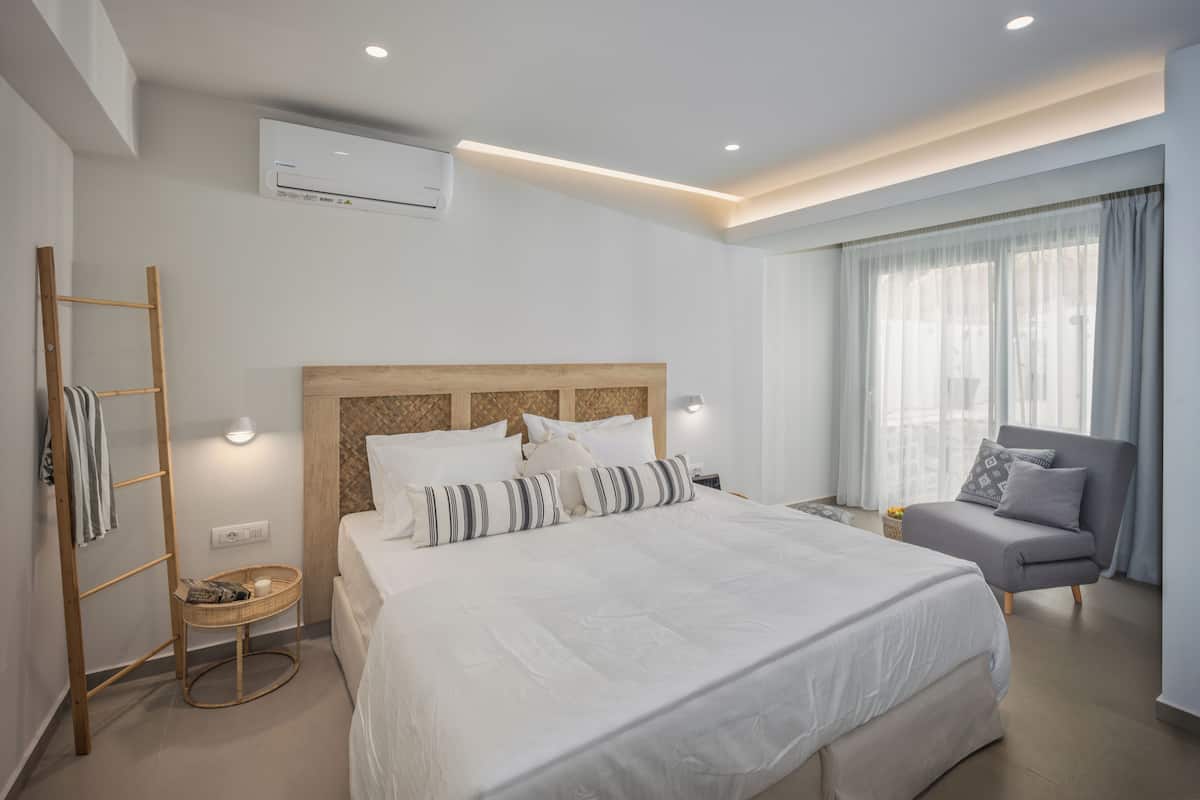 A modern hotel room with a large bed, a grey armchair, a small side table, and a window with sheer curtains.