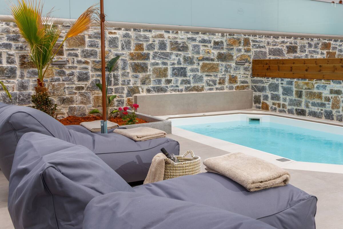 A poolside area with stone walls, a wooden deck, and grey lounge chairs.