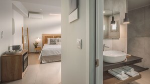 A modern hotel room with a large bed, a wooden bedside table, a built-in oven, a large wall-mounted mirror, and a large bathroom sink.