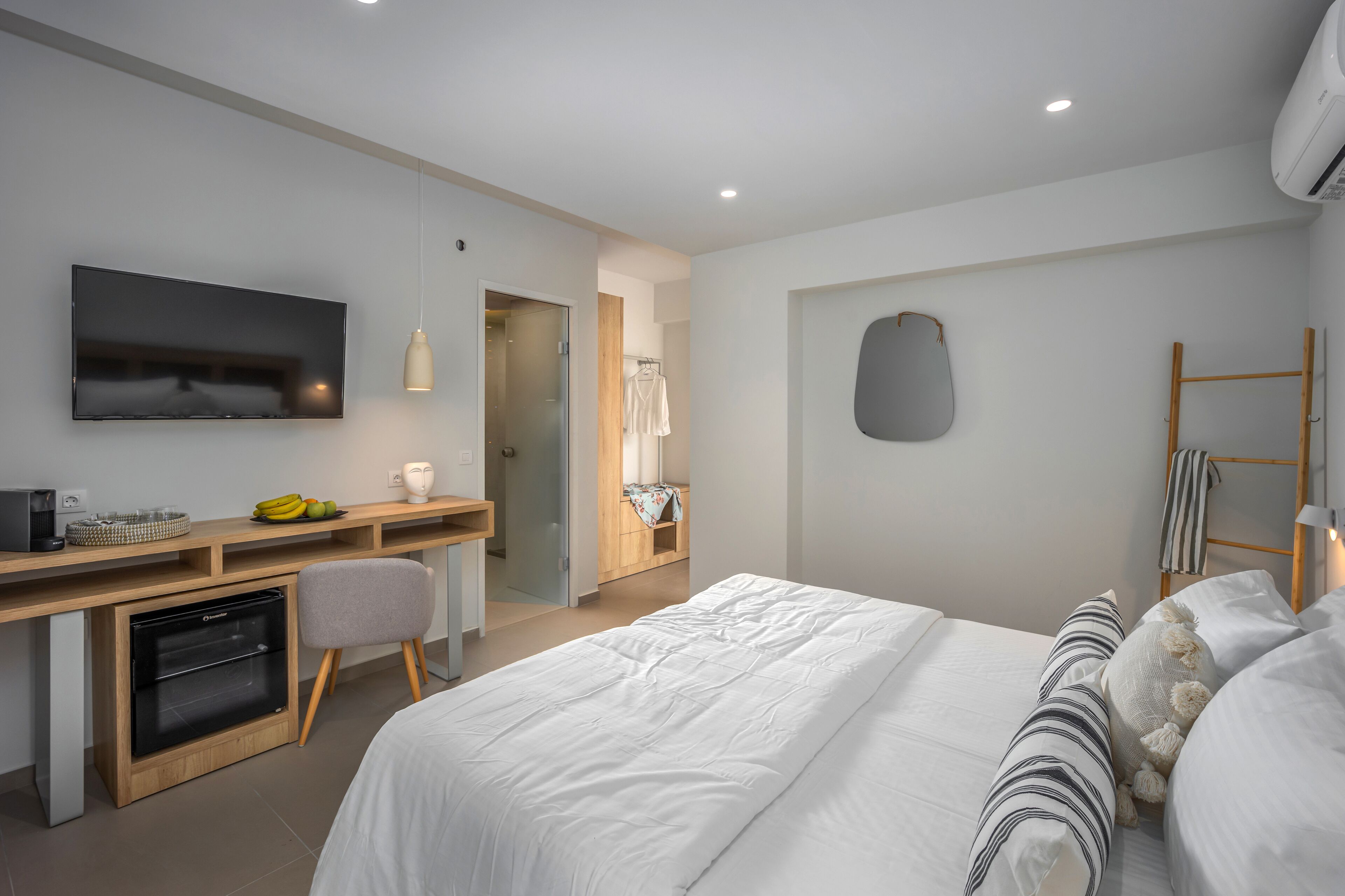 A modern hotel room with a bed, a desk with a computer, a television, and a small kitchenette.