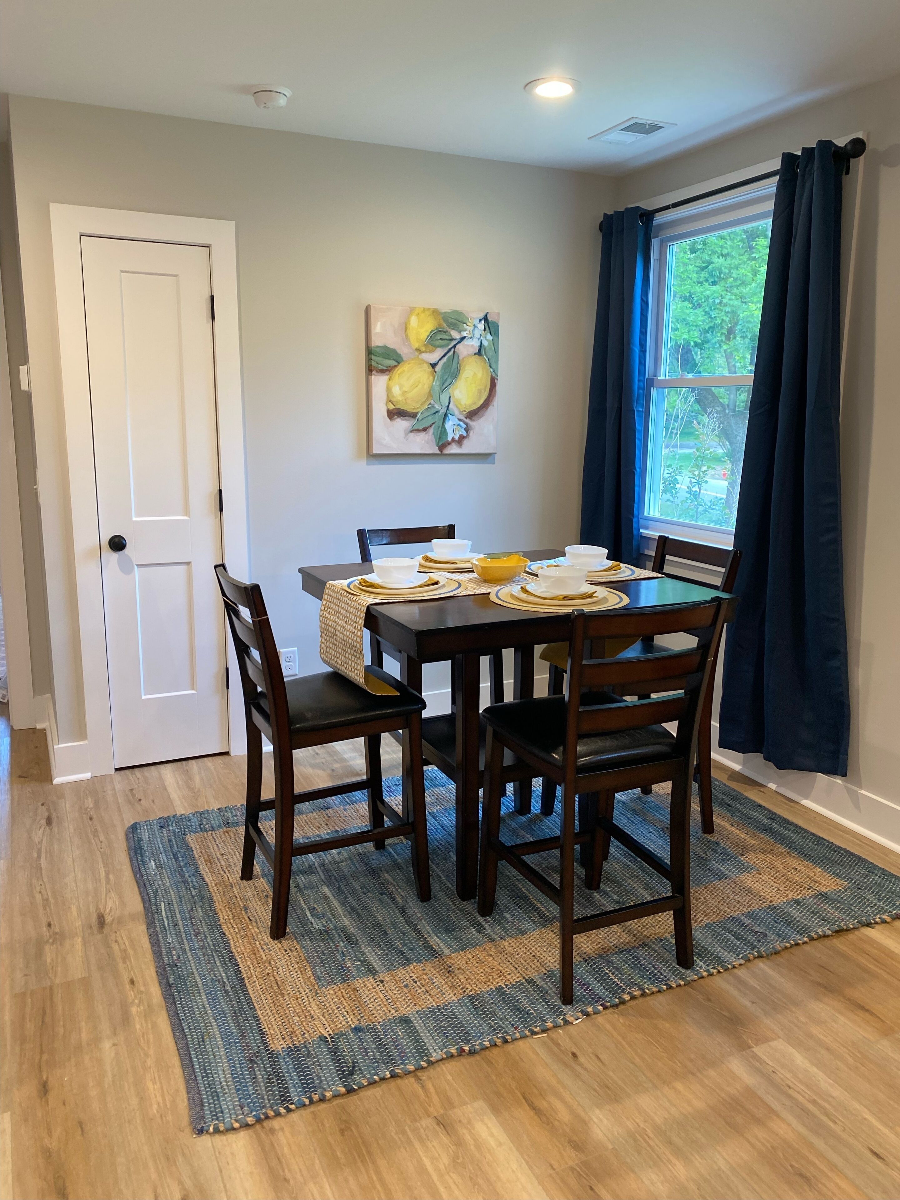 A dining area with a table set for four, a painting of lemons on the wall, and a window with curtains.