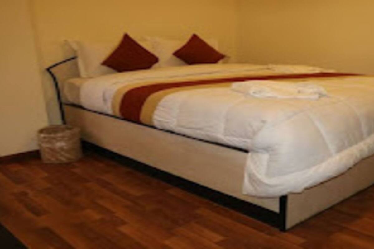 A single bed with a white duvet and two red pillows, a wooden floor, and a small trash bin.