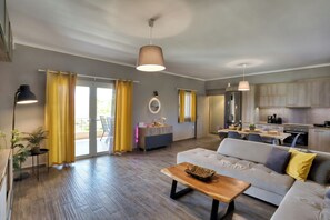 A spacious living area with a sofa, coffee table, and a dining table. There is a kitchen area with cabinets and a refrigerator. - Sitia Bay View Villa Apartment (Σητεία)