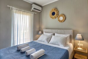 A hotel room with a bed, a bedside lamp, a wall-mounted air conditioner, and a window with sheer curtains. - Sitia Bay View Villa Apartment (Σητεία)