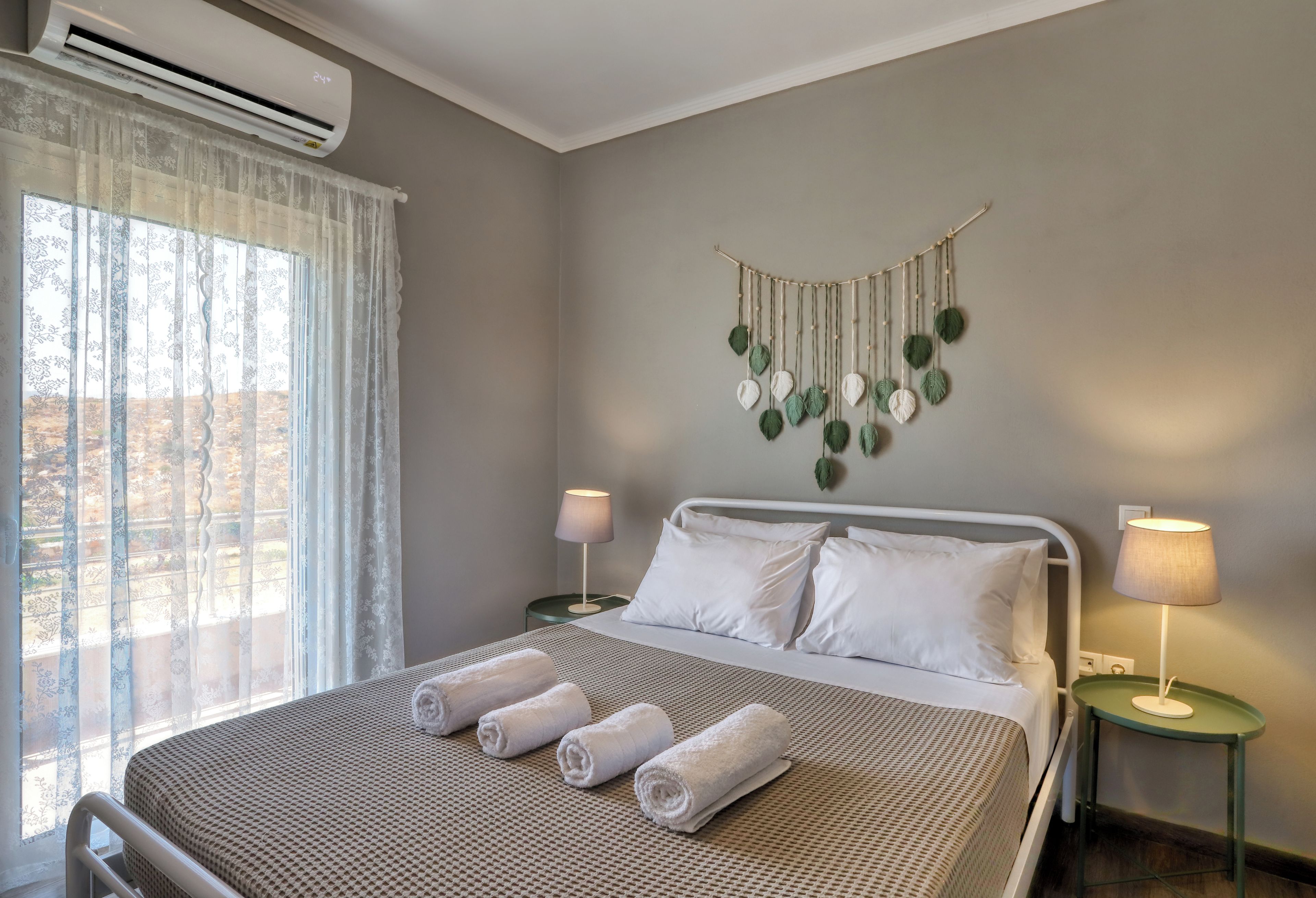 Ein Hotelzimmer mit einem Bett, zwei Nachttischlampen, einer Wanddekoration aus Makramee und einem Fenster mit durchsichtigen Vorhängen.