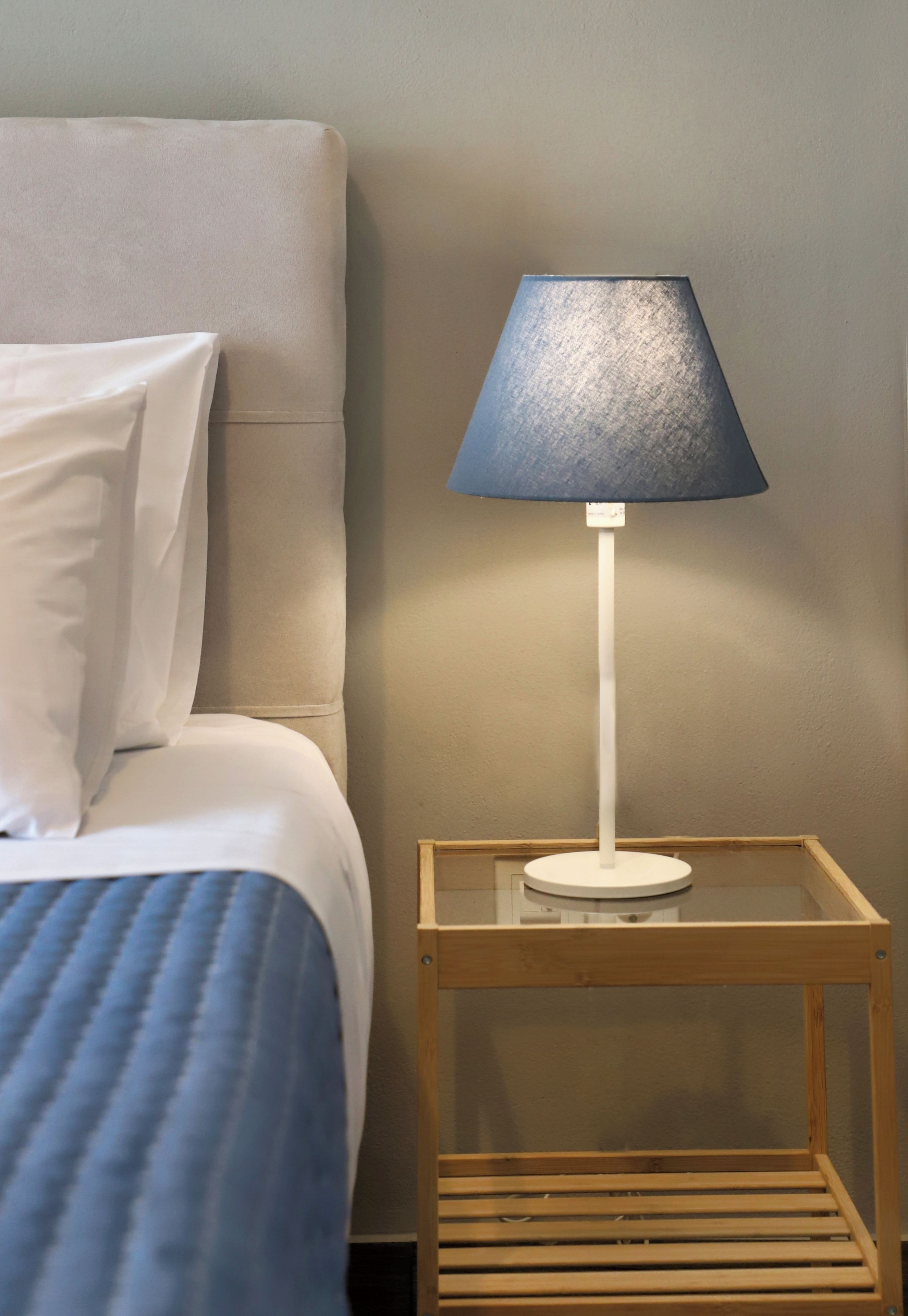 Een bed met witte beddengoed en een blauwe deken, een nachtkastje met een lamp, en een houten nachtkastje.