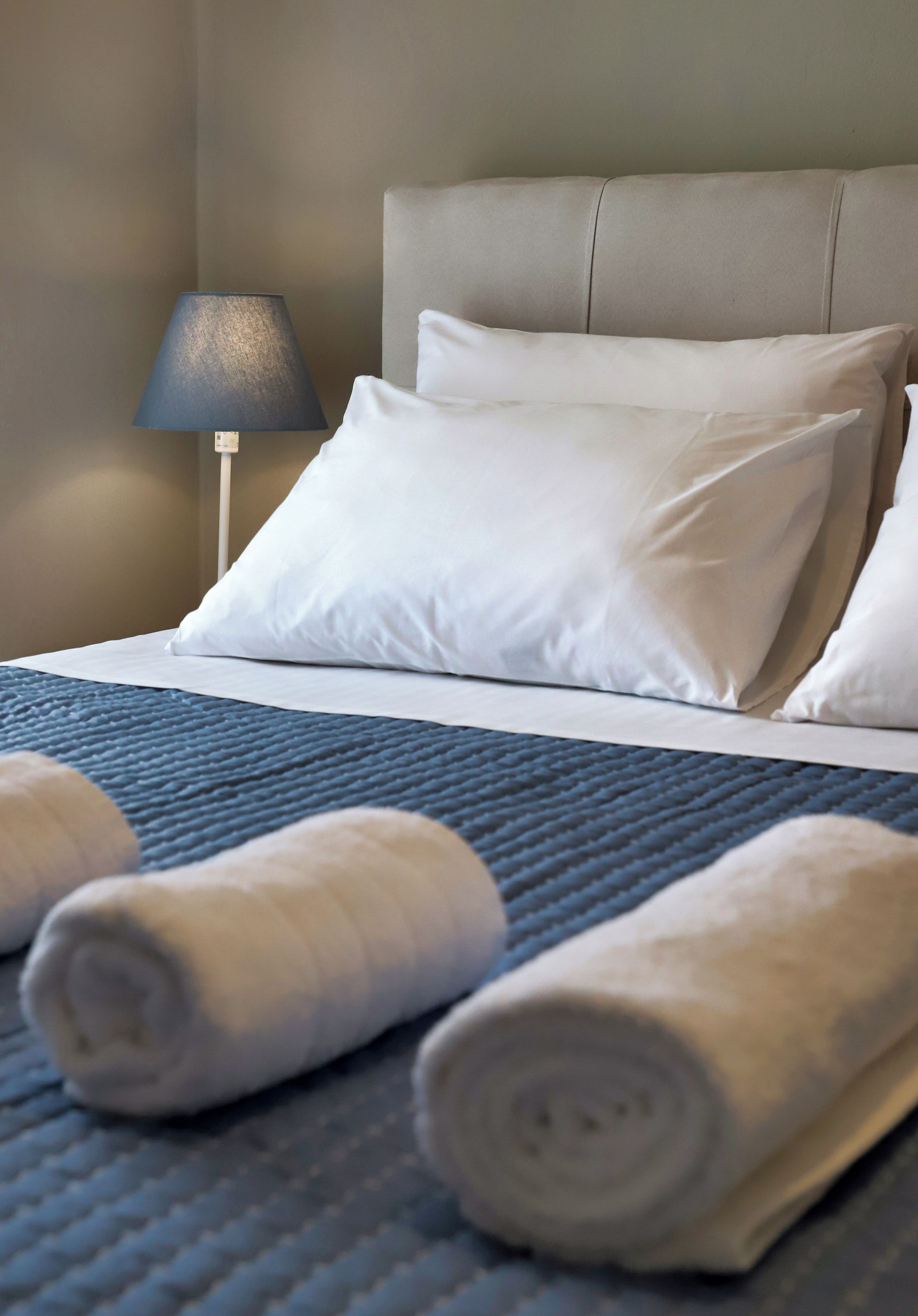Ein ordentlich bezogenes Bett mit weißen Kissen und einer blauen Bettdecke, dazu gerollte Handtücher.