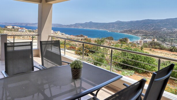A balcony with a table and chairs, overlooking a coastal landscape with mountains in the distance. - Sitia Bay View Villa Apartment (Σητεία)
