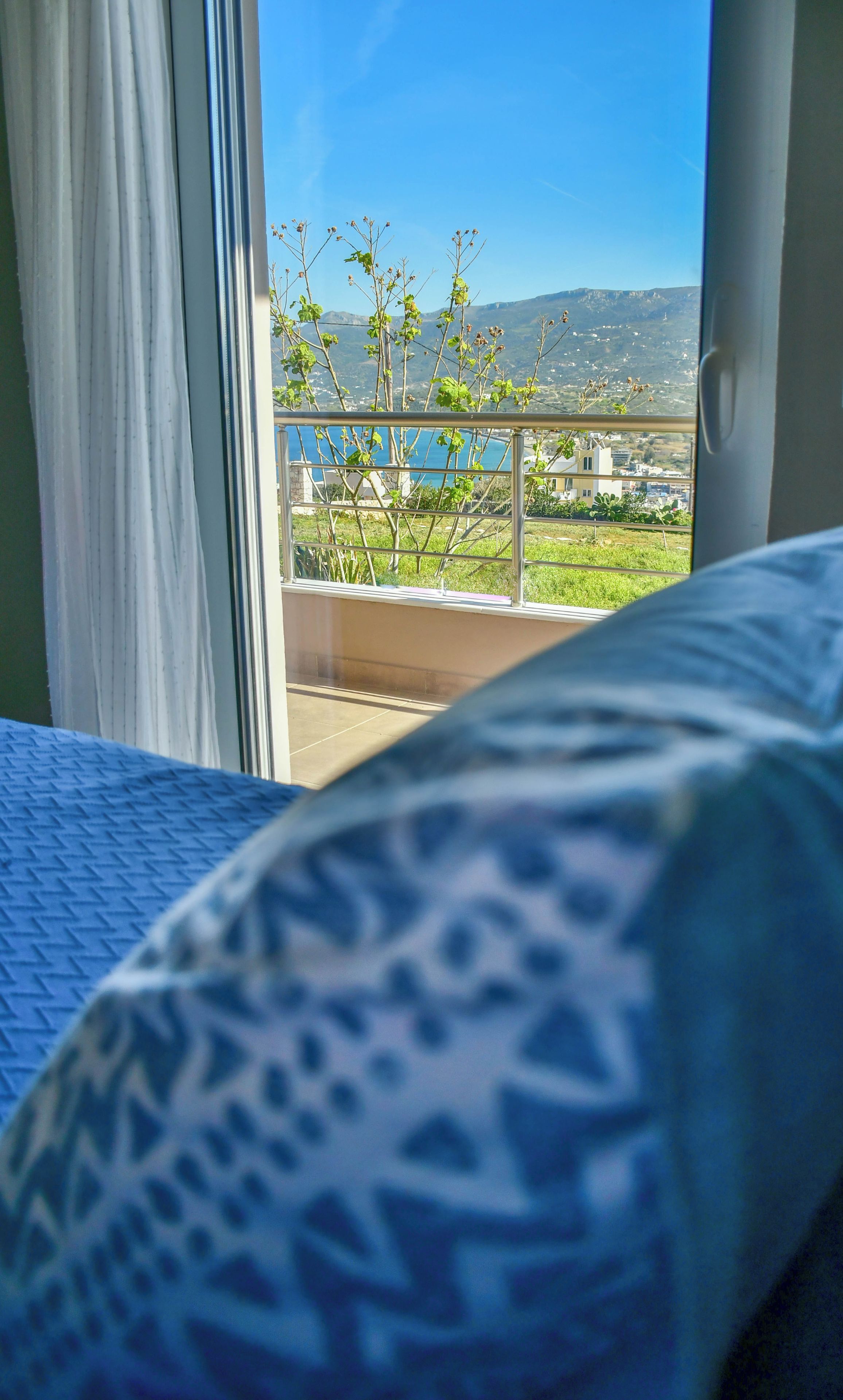 Quarto com cama, vista para uma varanda com plantas e paisagem distante.