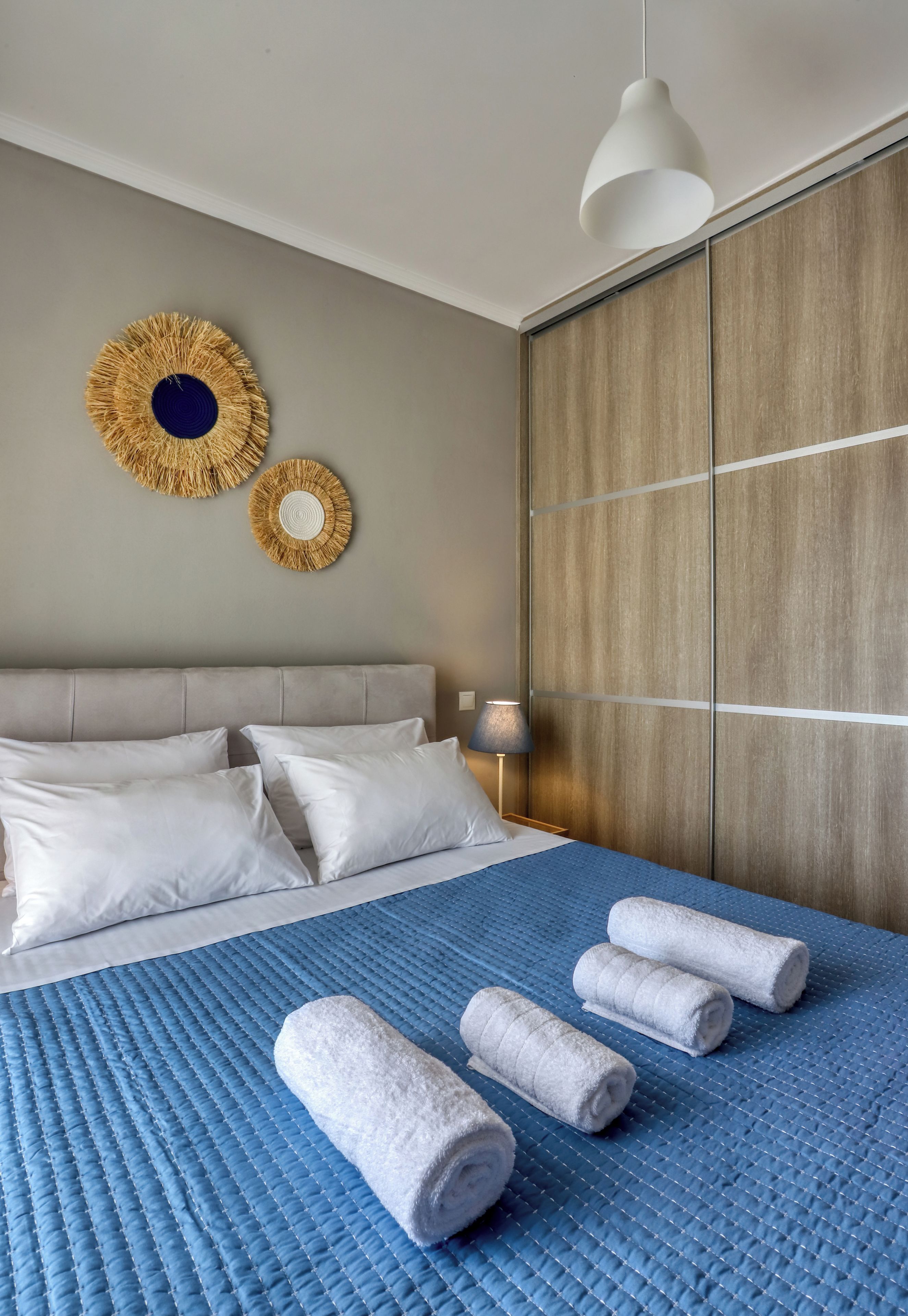 A hotel room with a bed, pillows, a blue quilted bedspread, two wall decorations, a hanging light fixture, and a wooden wardrobe.