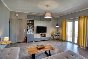 A modern living room with a flat-screen TV, wooden furniture, and a large window with yellow curtains. - Sitia Bay View Villa Apartment (Σητεία)