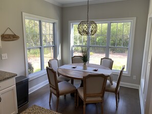 A dining area with a round table, six chairs, a hanging light fixture, and large windows with a view of greenery. - Coastal Retreat Minutes from the Beach/Oak Island and Historic Southport (Southport)