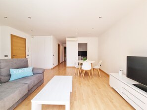 A modern living room with a grey sofa, a white coffee table, and a TV on a white cabinet. There is a dining area with a white table and chairs, and a kitchen in the background.
