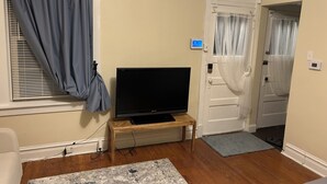 Una habitación con una televisión sobre un mueble de madera, una ventana con cortinas, una puerta con cortinaje y un termostato en la pared.