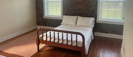 A bedroom with a wooden bed, two windows with blinds, a brick wall, and a wooden floor.