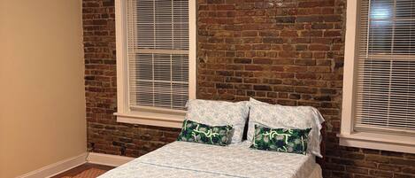 A bedroom with a wooden bed, two windows with blinds, a brick wall, and a ceiling light.