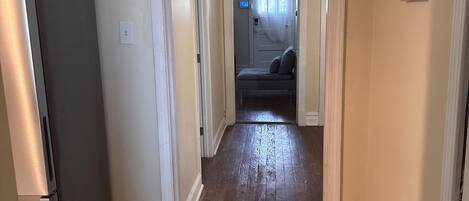 A hallway with a refrigerator on the left, a bench in the center, and a door leading to another room.