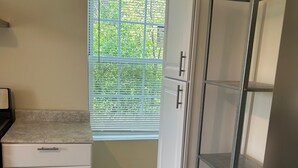 Una cocina con armarios blancos, una encimera, una ventana con persianas y una estantería metálica.