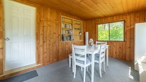 Una habitación con paneles de madera, una puerta blanca, una estantería, una mesa de comedor con sillas y un ventanal con vistas a árboles.