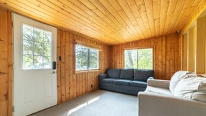 Interior de una cabaña de madera con una puerta blanca, dos ventanas, un sofá gris y un sillón beige.