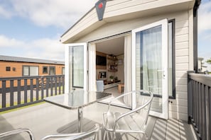 A modern RV with a glass door leading to a kitchen area, a patio with a table and chairs, and a wooden deck.