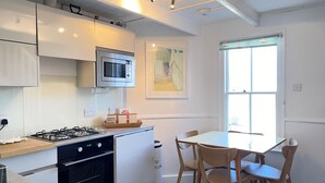 A compact kitchen with a gas stove, oven, microwave, and white cabinetry. There is a dining table with four chairs and a window with blinds.