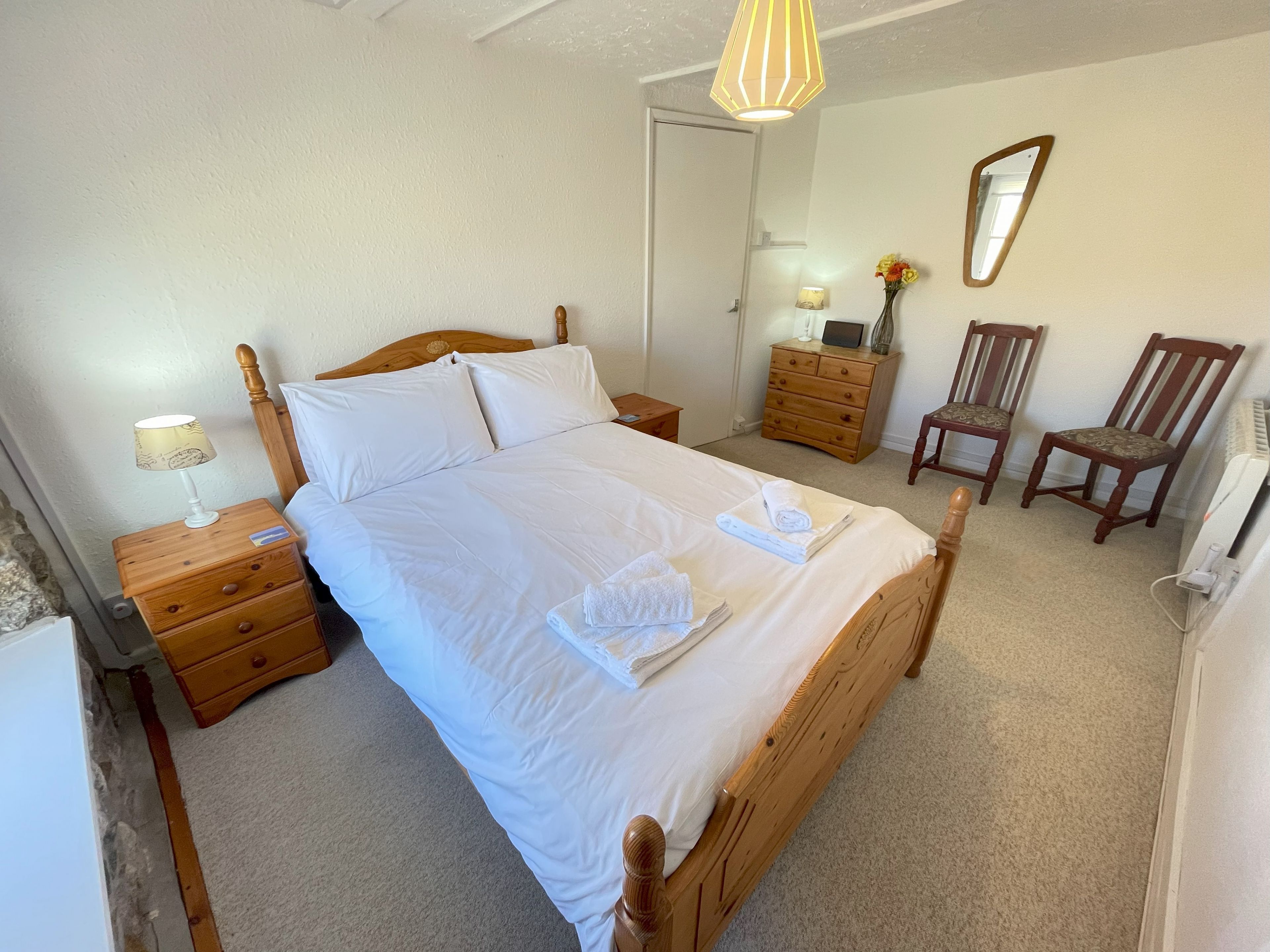 A neatly made bed with white linens and rolled towels, wooden nightstands with lamps, a dresser, and two wooden chairs.