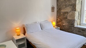 A bed with white bedding, two bedside lamps, a white nightstand, a stone wall, and a window with a view. - Lower Deck -  a cottage that sleeps 4 guests  in 2 bedrooms (St Ives)