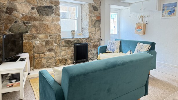 A cozy living room with a stone wall, a teal sofa, a white table with a flat-screen TV, and a hanging light fixture. - Lower Deck -  a cottage that sleeps 4 guests  in 2 bedrooms (St Ives)