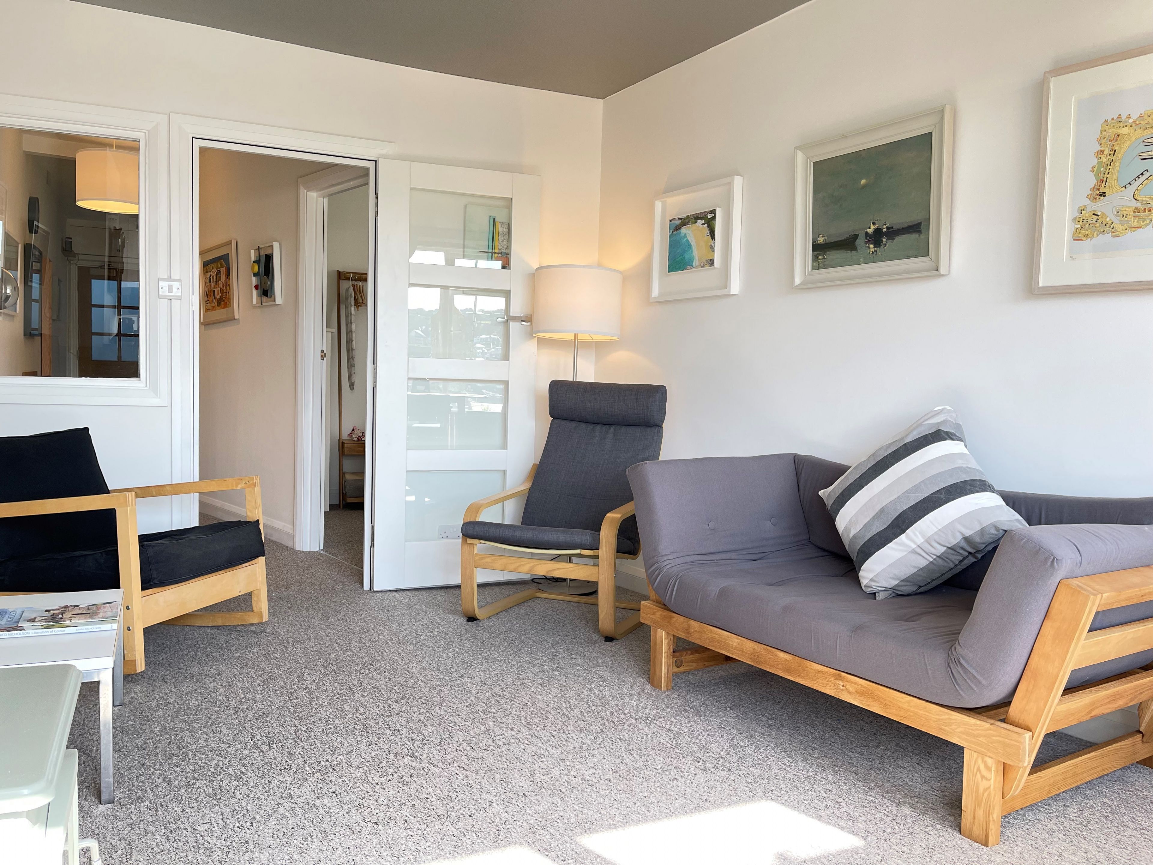 Ein Wohnzimmer mit grauem Sofa, einem Holzschaukelstuhl und einem kleinen Tisch. An der Wand befinden sich gerahmte Bilder und eine Tür, die in einen anderen Raum führt.