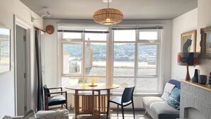 A cozy living room with a dining area, large windows with a view of the city, a fireplace, and a ceiling light fixture.