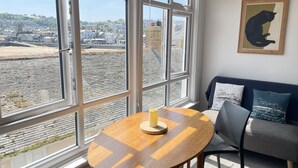 A room with large windows, a round wooden table with a candle, a grey sofa with patterned cushions, and a framed artwork on the wall.