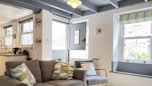 A cozy living room with a grey sofa, a wooden chair, and a window with a striped valance. - The Bolt Hole - a cottage that sleeps 4 guests in 2 bedrooms (St Ives)