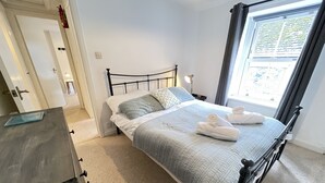 A neatly made bed with a quilted blanket and two pillows, a bedside lamp, and a window with curtains. - The Bolt Hole - a cottage that sleeps 4 guests in 2 bedrooms (St Ives)